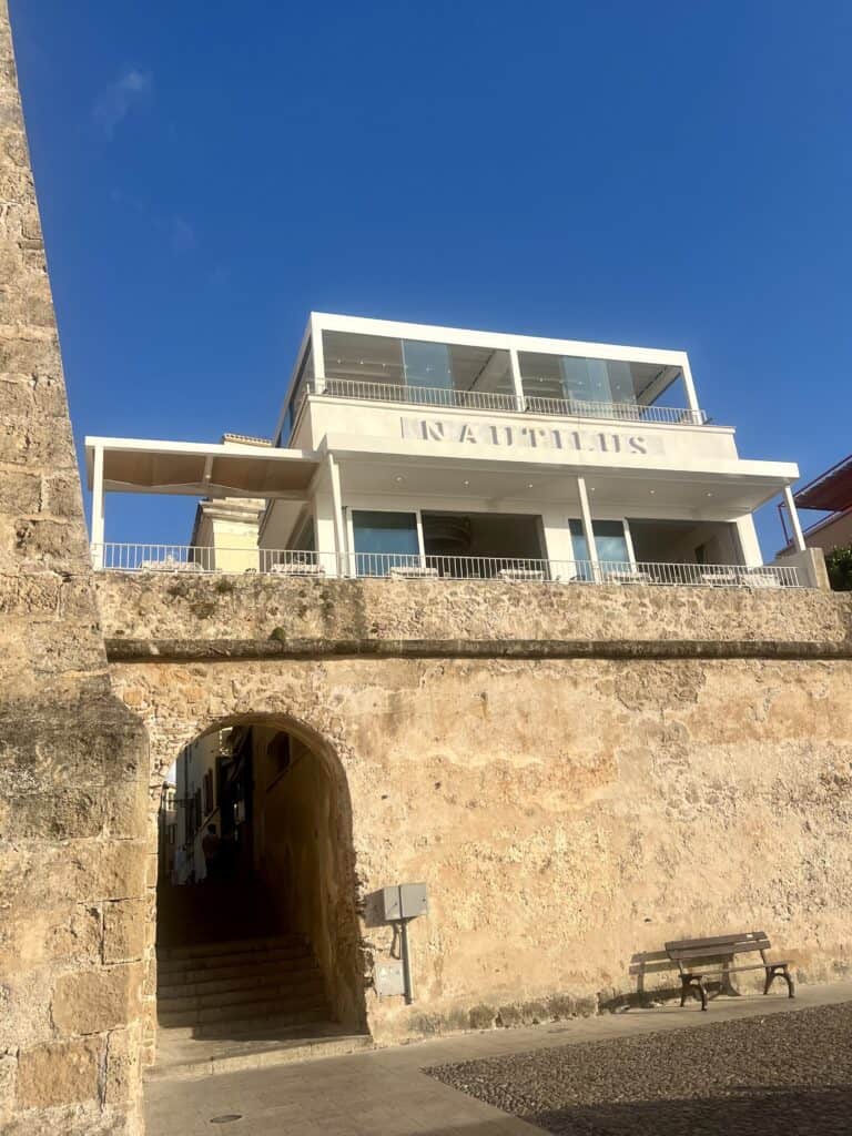 Modern white building with the word "Nautilus" above an arched passageway in a stone wall, under a clear blue sky—home to one of the best restaurants Alghero has to offer.