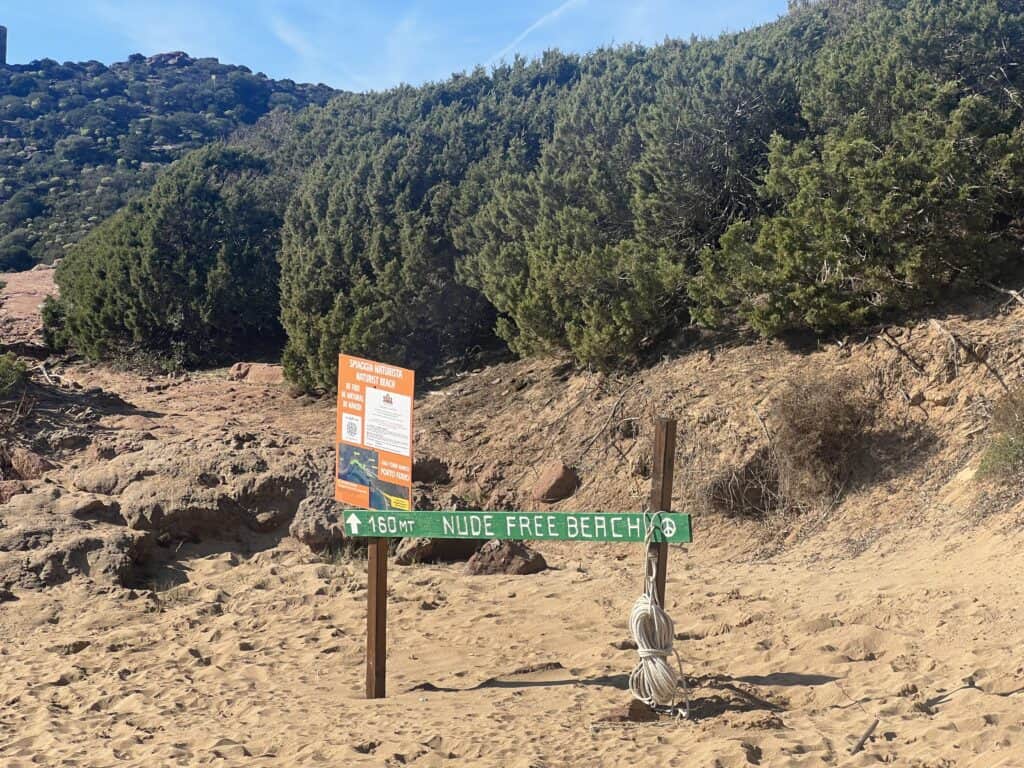 A wooden sign on a sandy path reads "Nude Free Beach" with an arrow and distance marker; among the best Alghero beaches, trees and informational signs are visible in the background.