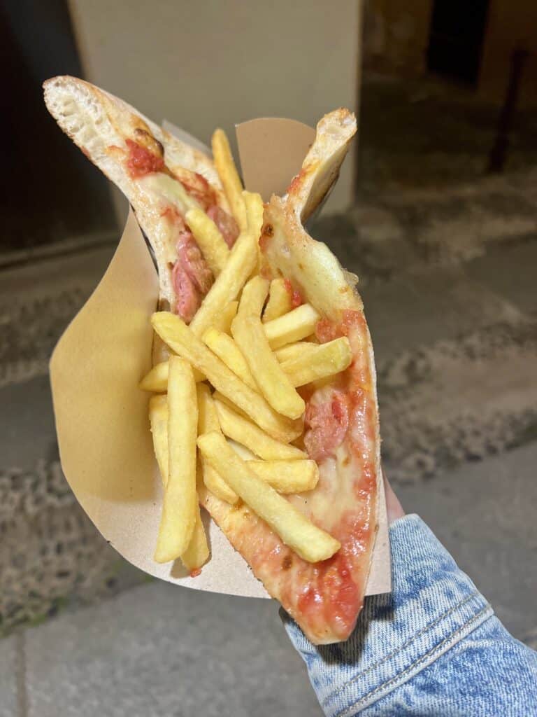 A hand holds a folded slice of pizza topped with French fries in a cardboard holder, set against a stone street—an experience you’ll find at some of the best restaurants Alghero has to offer.