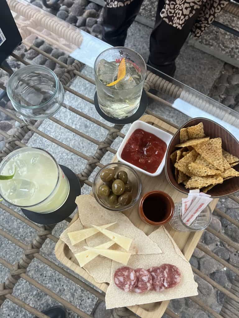 A table at one of the best restaurants Alghero has to offer, featuring two drinks, a bowl of nachos, green olives, red sauce, and a wooden board with cheese, crackers, and sliced salami.
