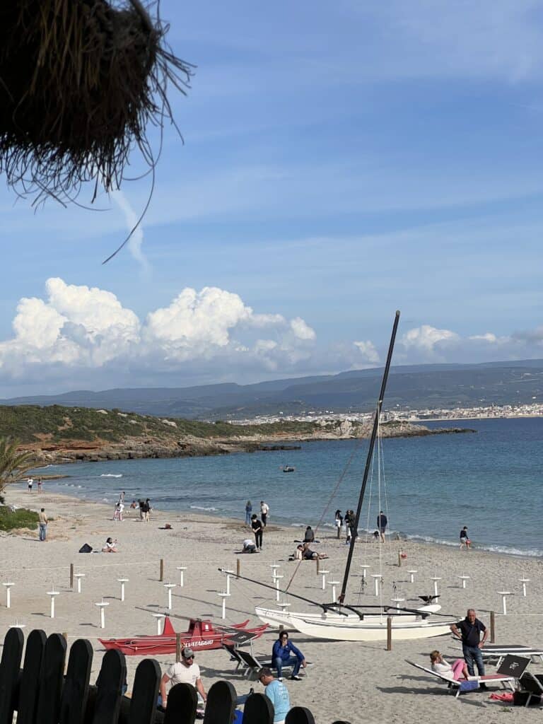 People are gathered on a sandy beach near the ocean, with sailboats on the shore and some people sitting or walking; distant hills and a town are visible in the background, capturing the charm of the best Alghero beaches.