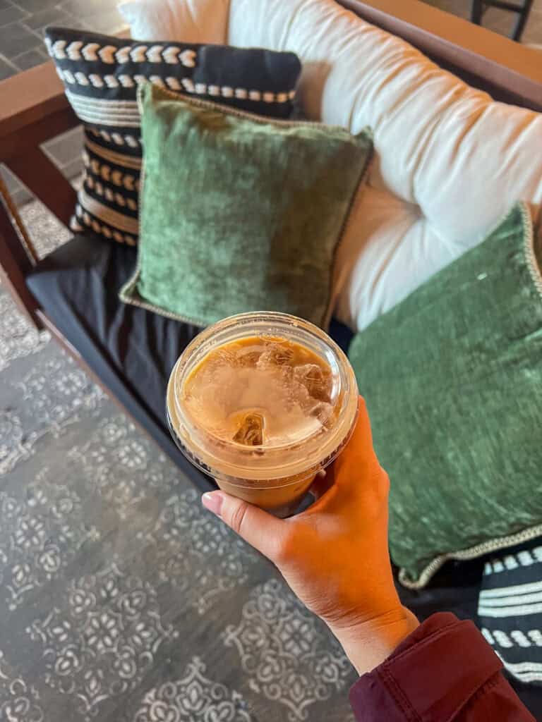 A hand holding an iced coffee in a plastic cup above a cushioned bench with green and black pillows, perfect for relaxing after reading western front hotel reviews.