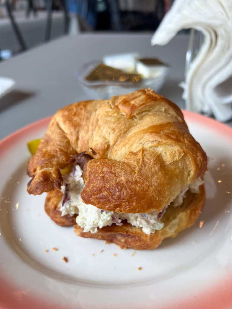 A croissant sandwich filled with creamy chicken salad sits on a white plate with a pink border; condiments and napkins are in the background, reminiscent of dishes praised in western front hotel reviews.