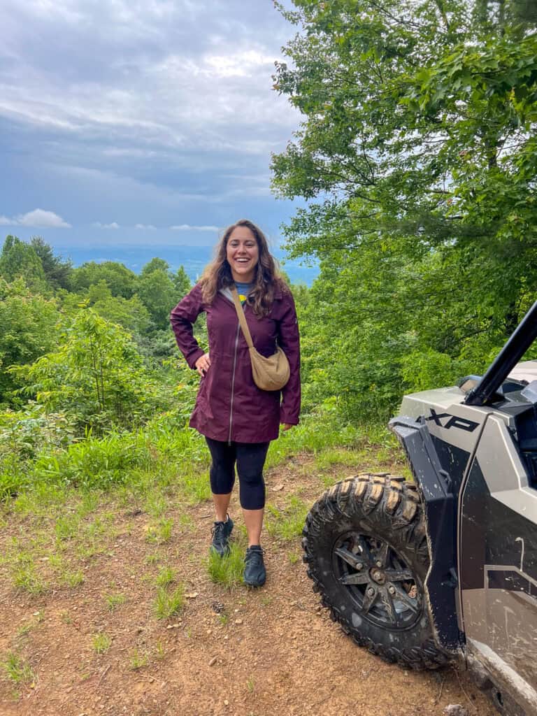 A woman in a maroon jacket stands on a dirt path beside an off-road vehicle, surrounded by green trees and an overcast sky, reminiscent of scenic spots featured in western front hotel reviews.