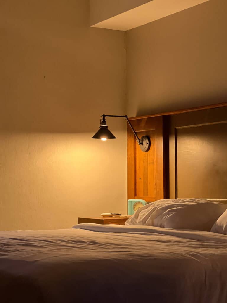 A neatly made bed with white linens is next to a wooden headboard and nightstand, softly lit by a wall-mounted reading lamp—an inviting scene often highlighted in Western Front Hotel reviews.