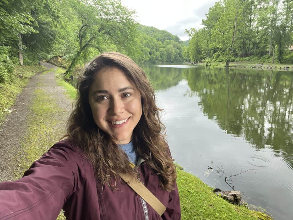 A woman with long brown hair takes a selfie by a calm river, surrounded by green trees and a walking path on a cloudy day, perhaps inspired by glowing western front hotel reviews.
