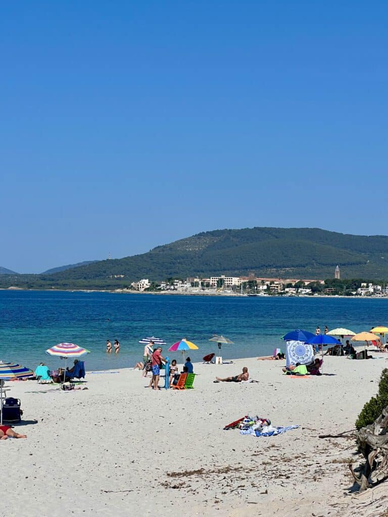 People relax under colorful umbrellas on a sandy beach with clear blue water and hills in the background—a perfect scene from the best Alghero beaches on a sunny day.