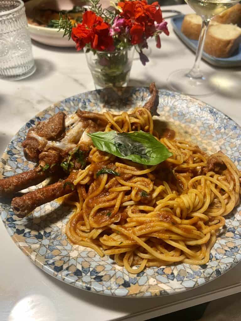 A plate of spaghetti with tomato sauce and soft-shell crab, garnished with basil, served on a patterned plate—just one of the delicious dishes you’ll find at the best restaurants Alghero has to offer. Bread, wine, water, and flowers complete the scene.