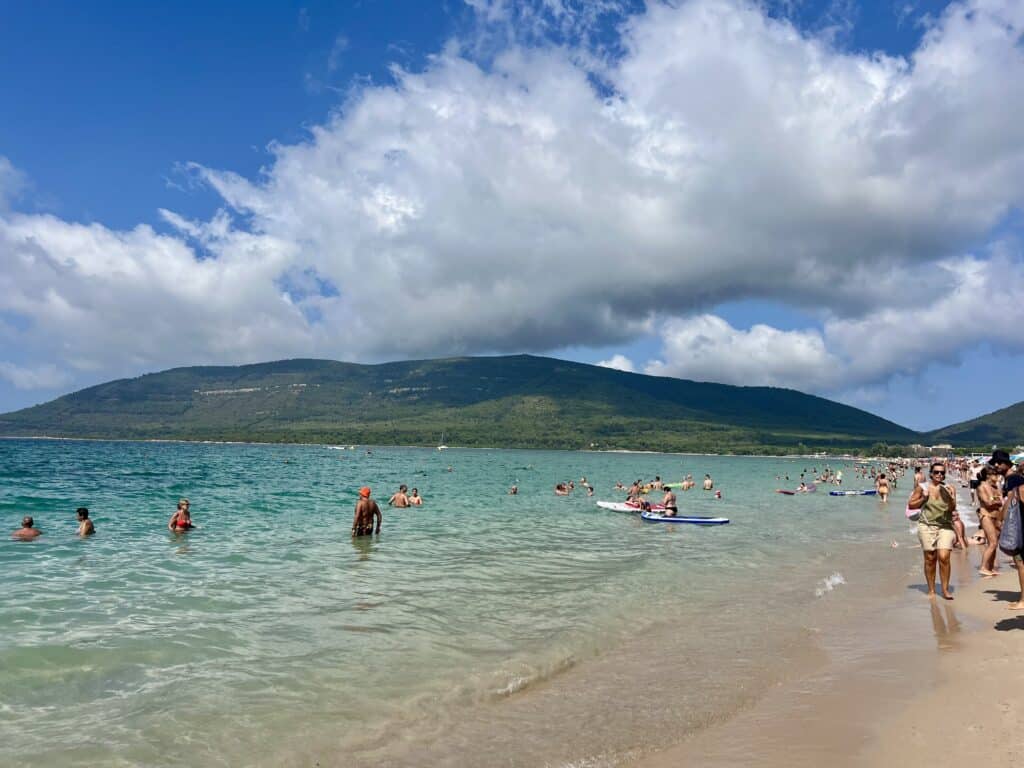 People swimming and relaxing on a sandy beach with clear water, grassy hills, and a partly cloudy sky in the background—one of the best Alghero beaches for an unforgettable coastal escape.