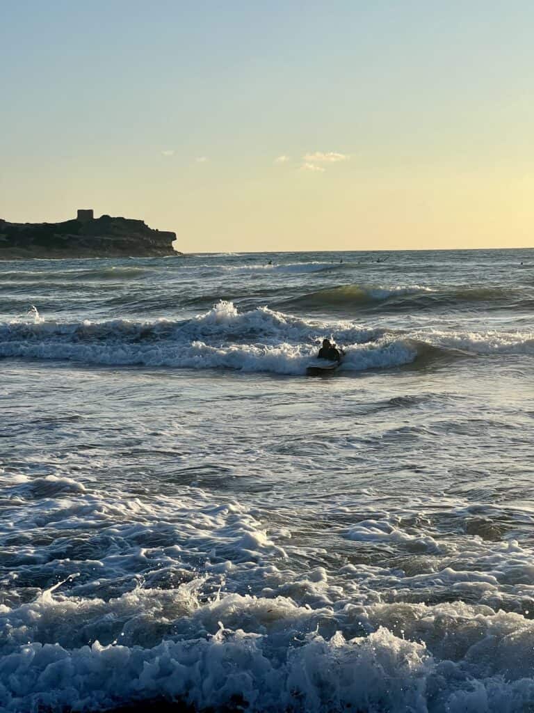 A person surfs on small waves near the shore at one of the best Alghero beaches, with a rocky headland and a building visible in the background under a clear sky.