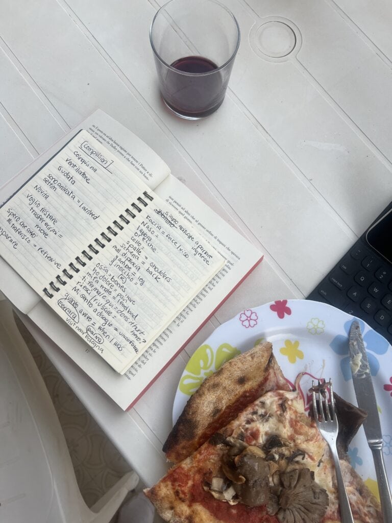 A notebook with handwritten notes, a plate with pizza slices, a fork and knife, a glass of red drink, and part of a tablet—perfect for planning visits to the best restaurants Alghero has to offer—all on a white table.