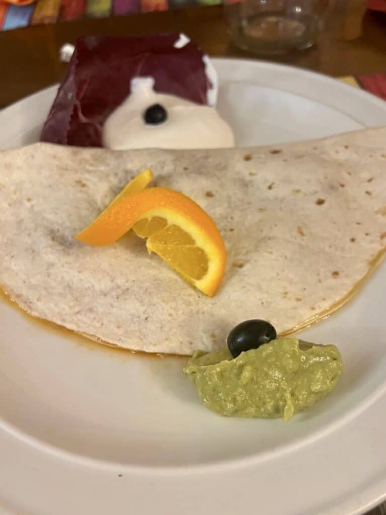 A folded flour tortilla on a white plate, garnished with an orange slice, guacamole, and a black olive; dessert with whipped cream and a single blueberry in the background—dishes you'd expect at the best restaurants Alghero has to offer.
