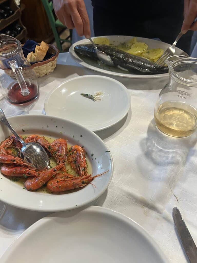 A table at one of the best restaurants Alghero features a plate of cooked shrimp, a platter of whole fish with potatoes, a basket of bread, and glass pitchers of white and red wine.