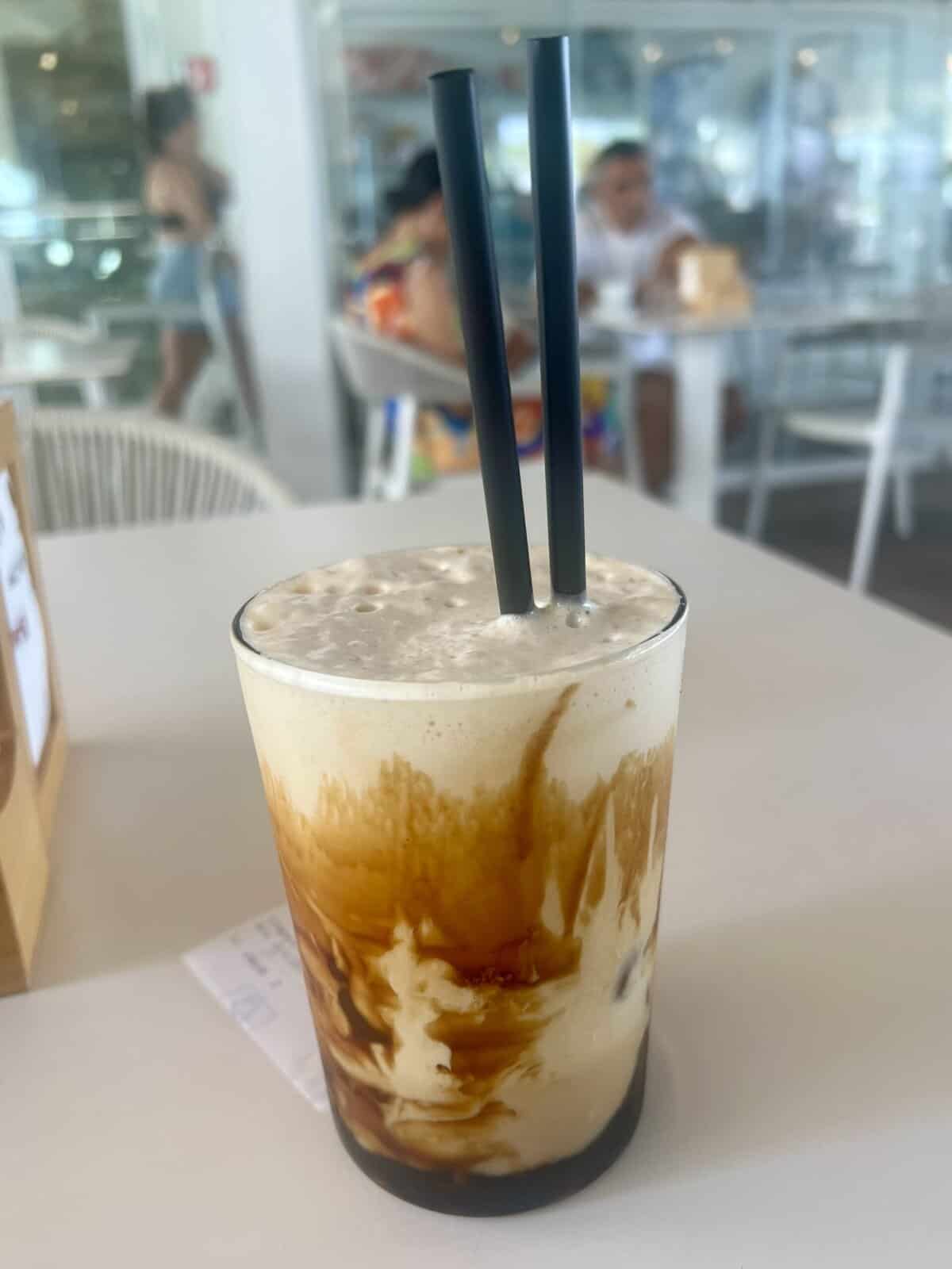 A glass of iced coffee with frothy milk and two black straws sits on a white table in a cafe, capturing the relaxed vibe often found in the best restaurants Alghero has to offer, with people and tables in the background.