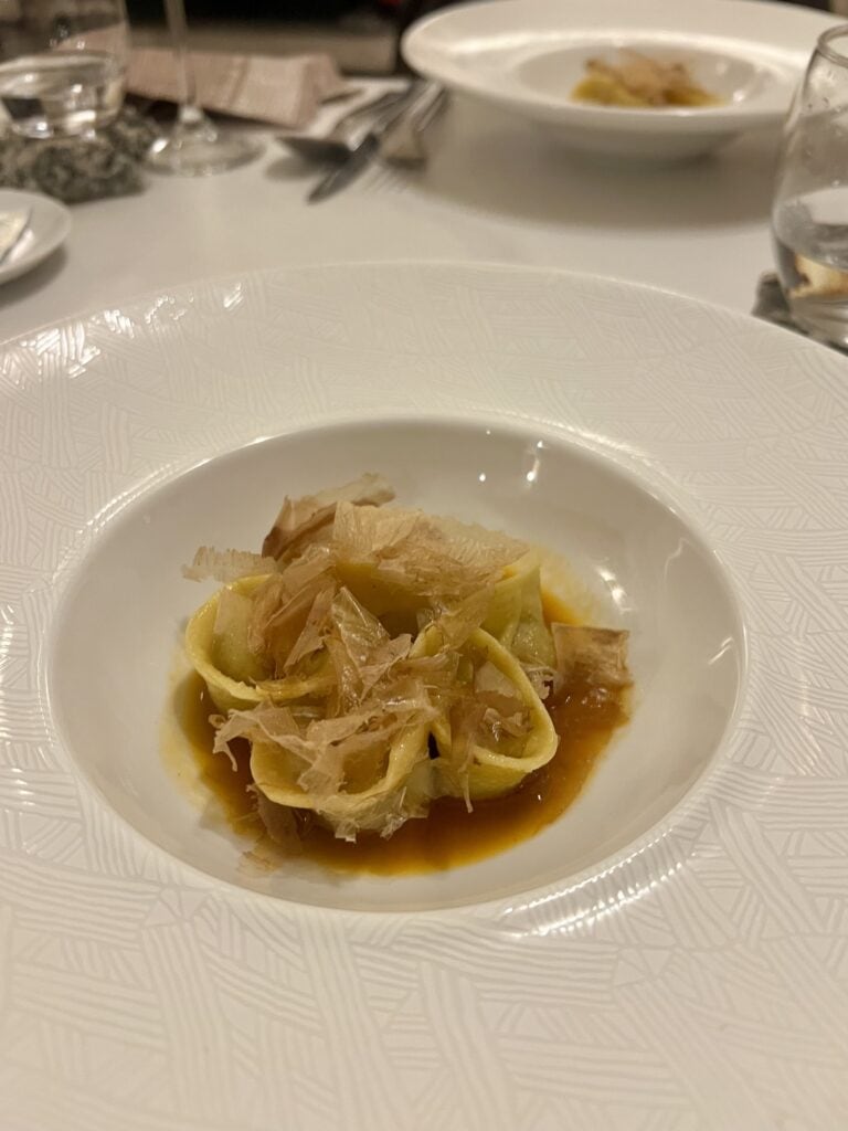 A white bowl contains pasta topped with thin flakes in a brown sauce, set on a white tablecloth—an inviting scene you might find at the best restaurants Alghero has to offer. Glassware and another plate can be seen in the background.