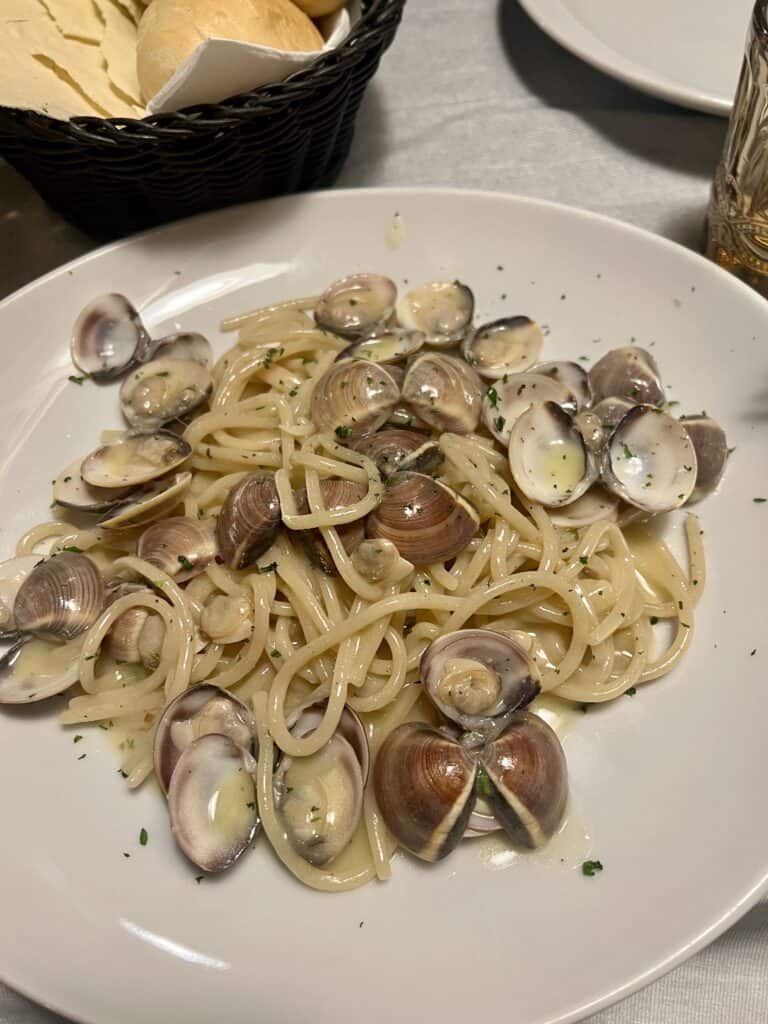 A plate of spaghetti with clams in a light sauce is served on a white dish, evoking the coastal flavors found in the best restaurants Alghero offers, with a bread basket and a glass visible in the background.