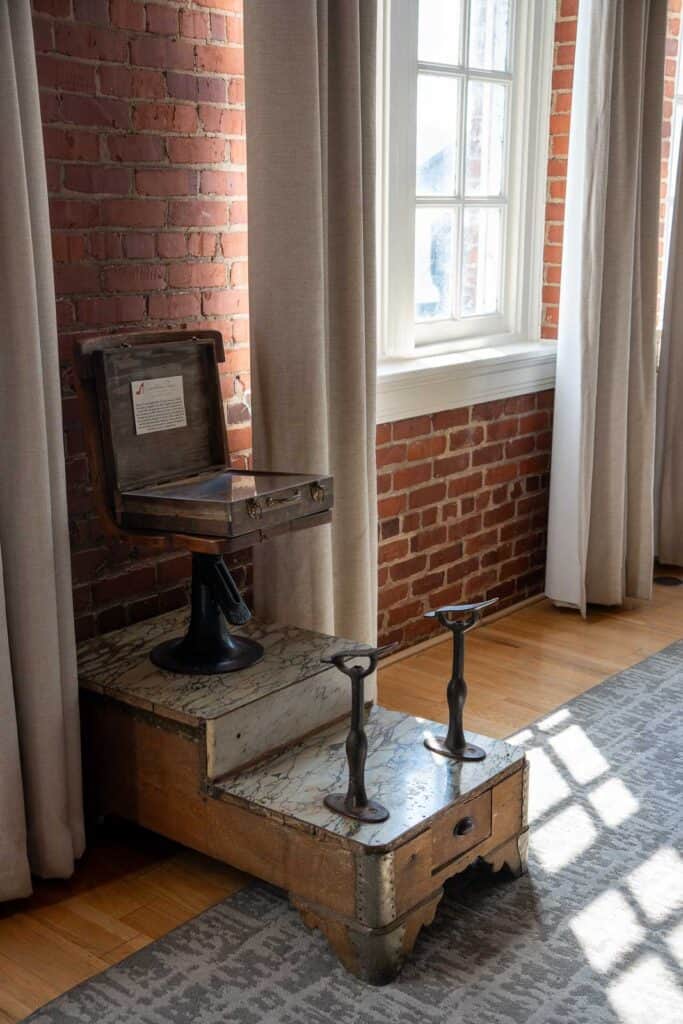 An old-fashioned wooden barber's chair with metal footrests sits on a small platform by a window in a brick-walled room at one of the best downtown Lynchburg hotels, featuring beige curtains and a patterned rug.