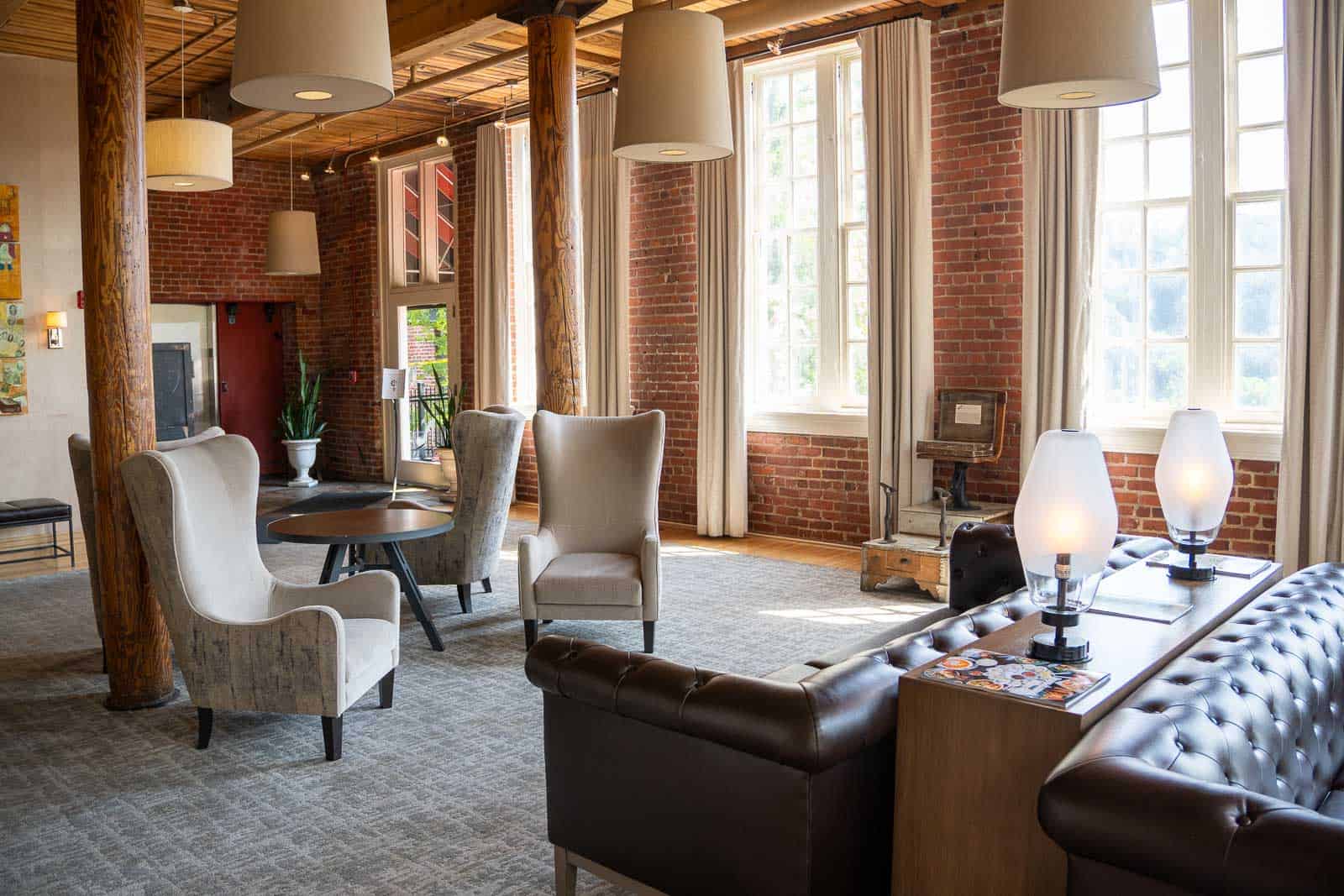 A spacious lounge at the Craddock Terry Hotel features exposed brick walls, tall windows, wooden beams, armchairs, a sofa, round tables, and modern lamps. Natural light fills the room in downtown Lynchburg.