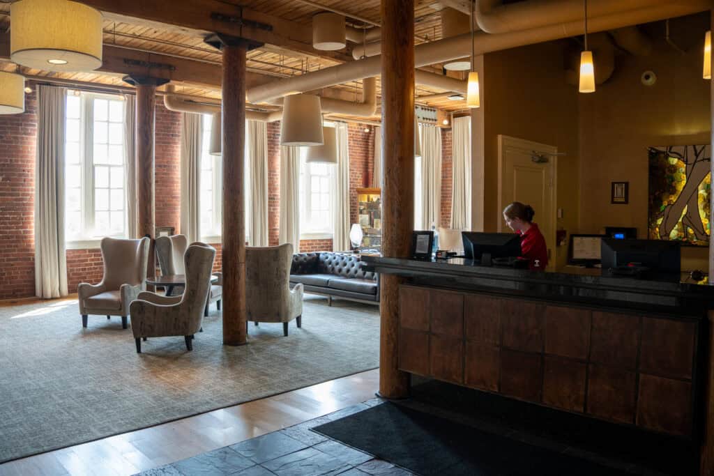 A hotel lobby with large windows, exposed brick walls, wooden beams, and a cozy seating area welcomes you—perfect for those seeking authentic downtown Lynchburg hotels. A receptionist is ready to assist at the front desk.