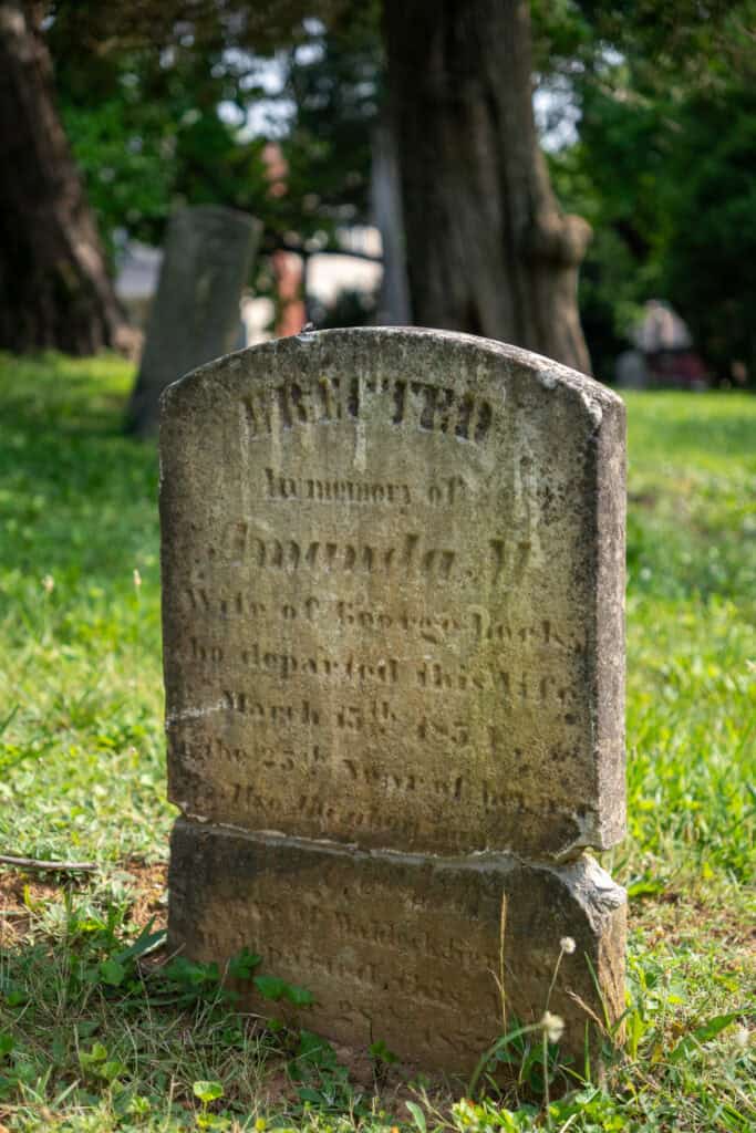 Weathered gravestone in a grassy cemetery, partially legible with the name Amanda, surrounded by trees and another headstone in the background—a tranquil scene far removed from downtown Lynchburg hotels.