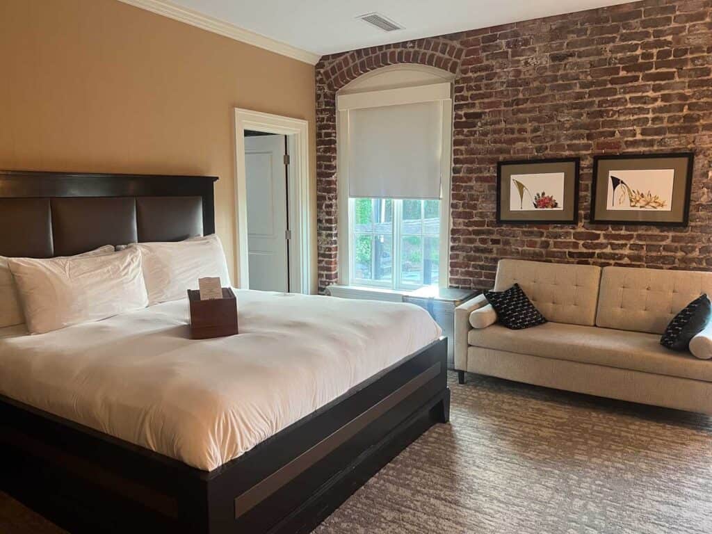 A bedroom at one of the best downtown Lynchburg hotels, featuring an exposed brick wall, a large bed with white linens, a beige sofa, two framed floral pictures, and a window with a white shade. Perfect for your next Craddock Terry Hotel review.