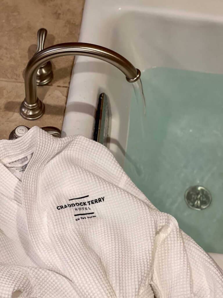 A white Craddock Terry Hotel robe is draped over the edge of a bathtub as water fills the tub from a silver faucet, reflecting the luxurious comfort found in downtown Lynchburg hotels.