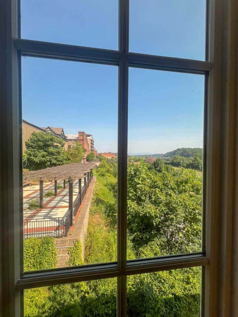 A view through a window showing a terrace with a pergola, lush green vegetation, buildings on the left, and a clear blue sky—perfect for those seeking downtown Lynchburg hotels with scenic charm.