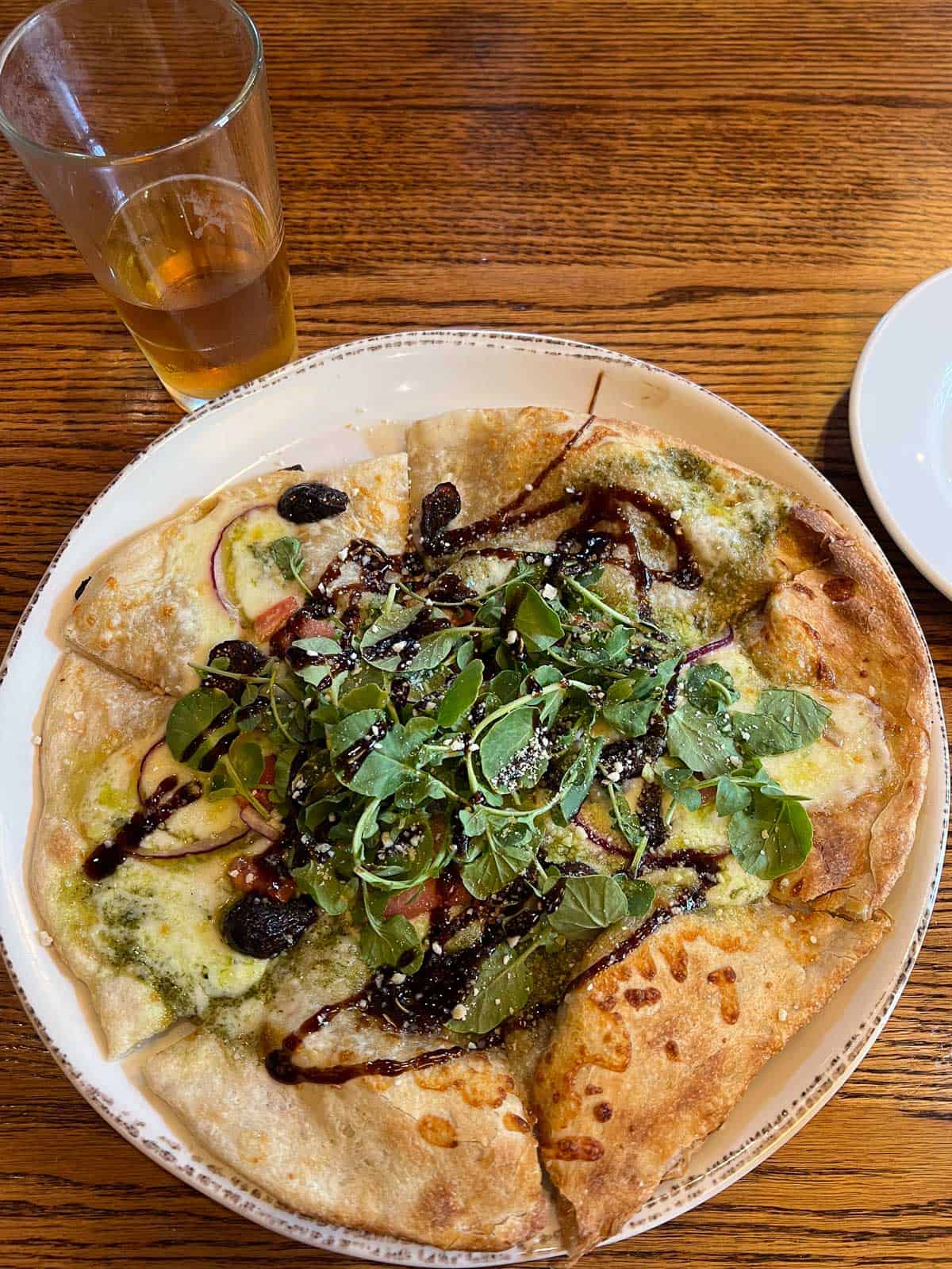 A sliced pizza topped with greens, balsamic glaze, and cheese sits on a plate next to a glass of beer on a wooden table—perfect for enjoying after reading a Craddock Terry Hotel review or exploring downtown Lynchburg hotels.