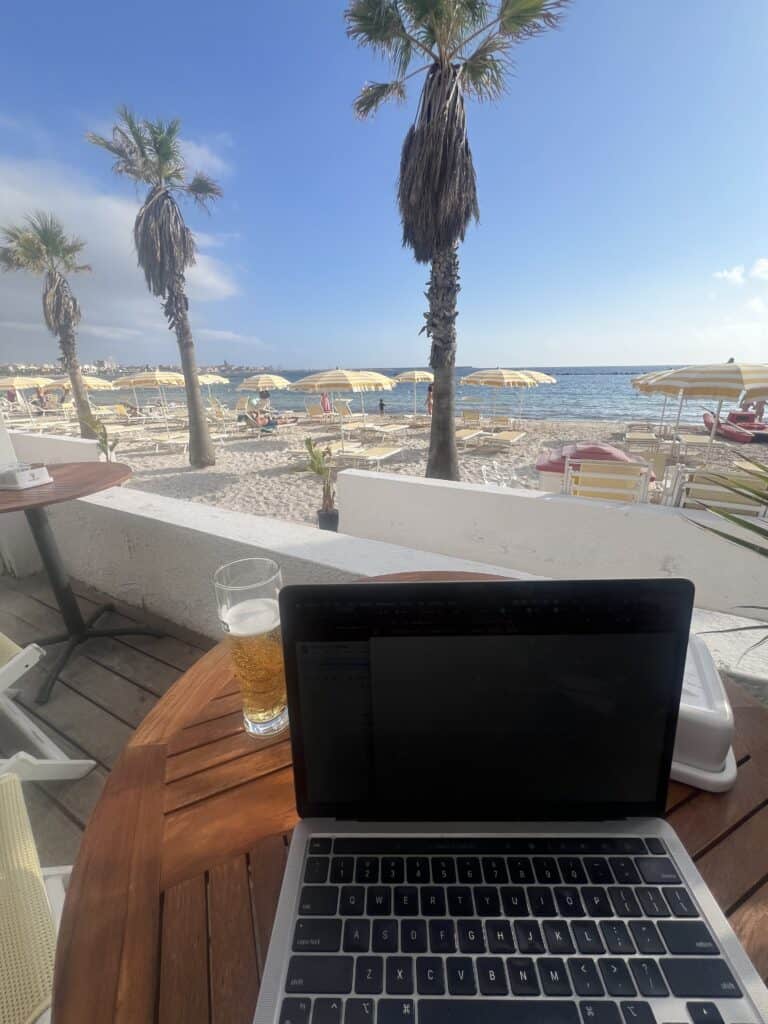A laptop and a glass of drink sit on a wooden table overlooking a sandy beach with palm trees and yellow umbrellas—perfect for browsing with an eSIM for Europe under a partly cloudy sky.