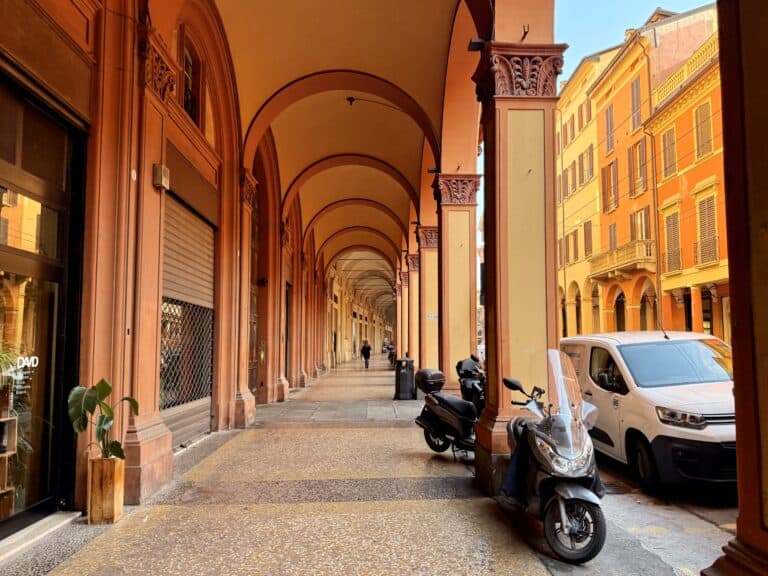 A covered walkway with columns and arches lines a city street. Parked scooters and cars are on the right, stores and closed shops are on the left, with a person walking in the distance.
