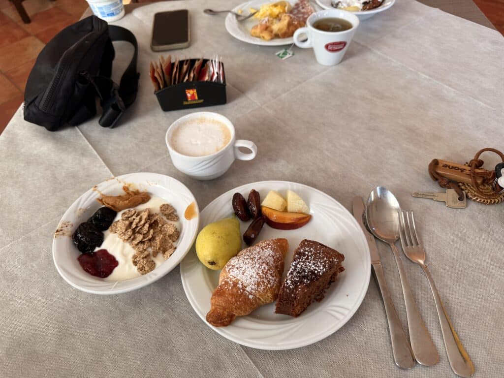 A breakfast table with plates of pastries, fruit, yogurt with granola and jam, coffee, utensils, a black bag, keys, and sugar packets—fuel for your Gorropu Gorge hike before exploring the stunning Su Gorropu Canyon.
