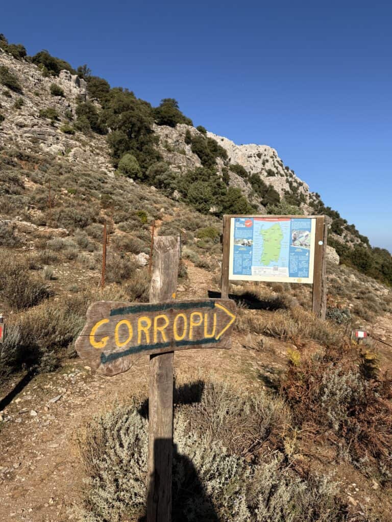 A wooden sign labeled "GORROPU" points toward the trail for the Gorropu Gorge hike, with a large informational map and rocky hills in the background under a clear blue sky.
