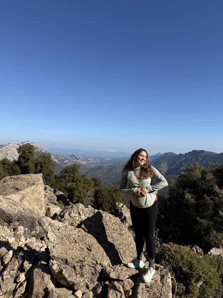 A person stands on rocky terrain with trees and distant mountains under a clear blue sky, capturing the rugged beauty of the Gorropu Canyon.
