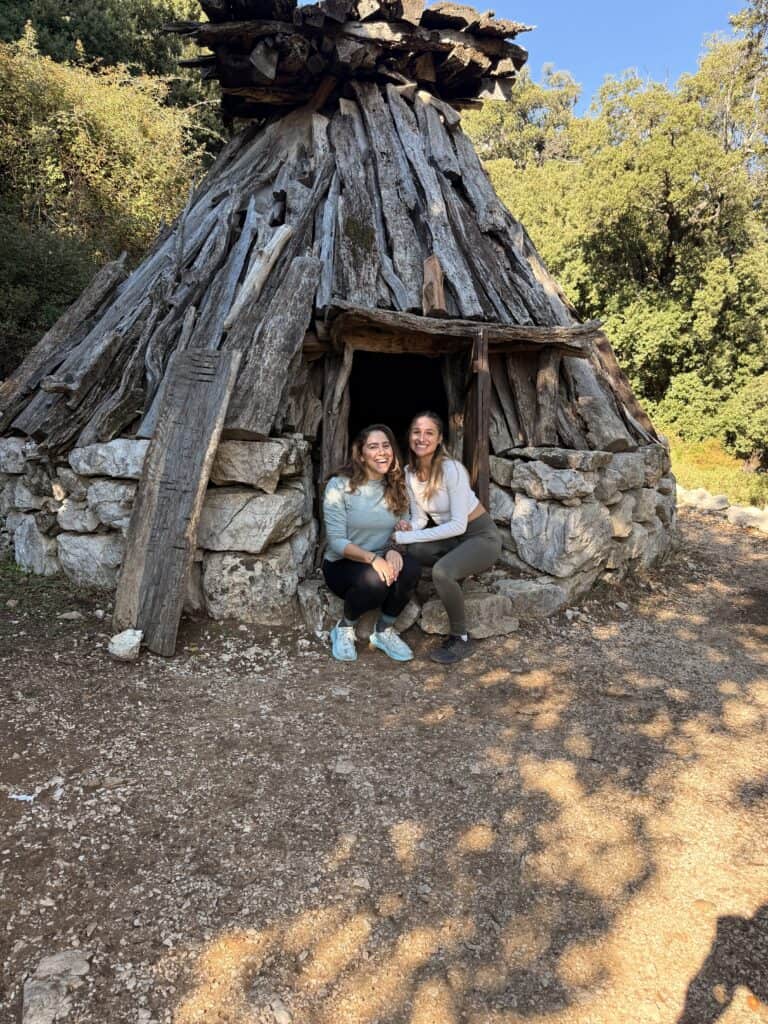 Two women are sitting and smiling in front of a small, rustic wooden and stone shelter surrounded by trees, taking a break during their Gorropu Gorge hike.