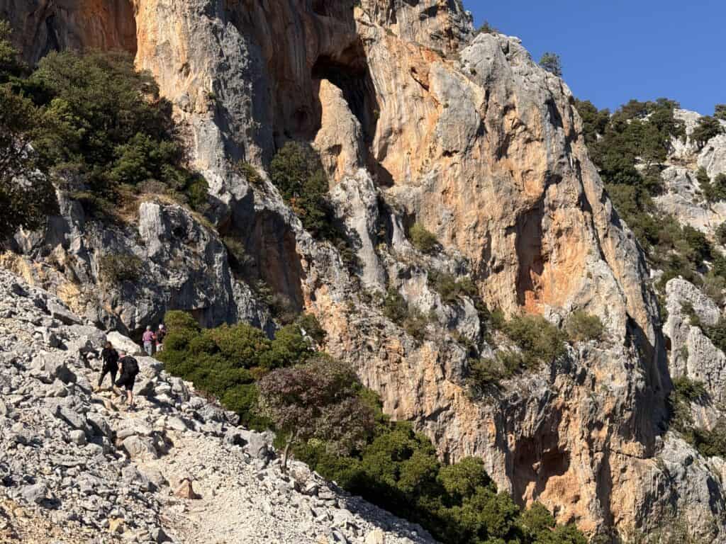 Several people hike along a rocky trail at the base of a steep, rugged cliff in Gorropu Canyon, with patches of green vegetation beneath a clear blue sky.