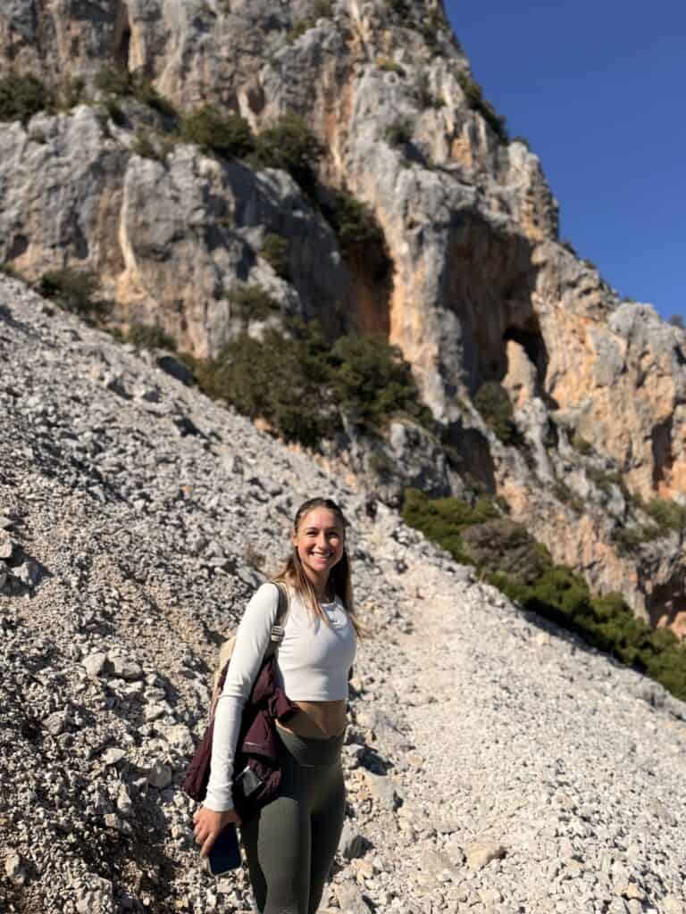 A woman stands on a rocky slope at Su Gorropu Canyon in front of a sunlit cliff under a clear blue sky, wearing athletic clothing and smiling at the camera after her Gorropu Gorge hike.
