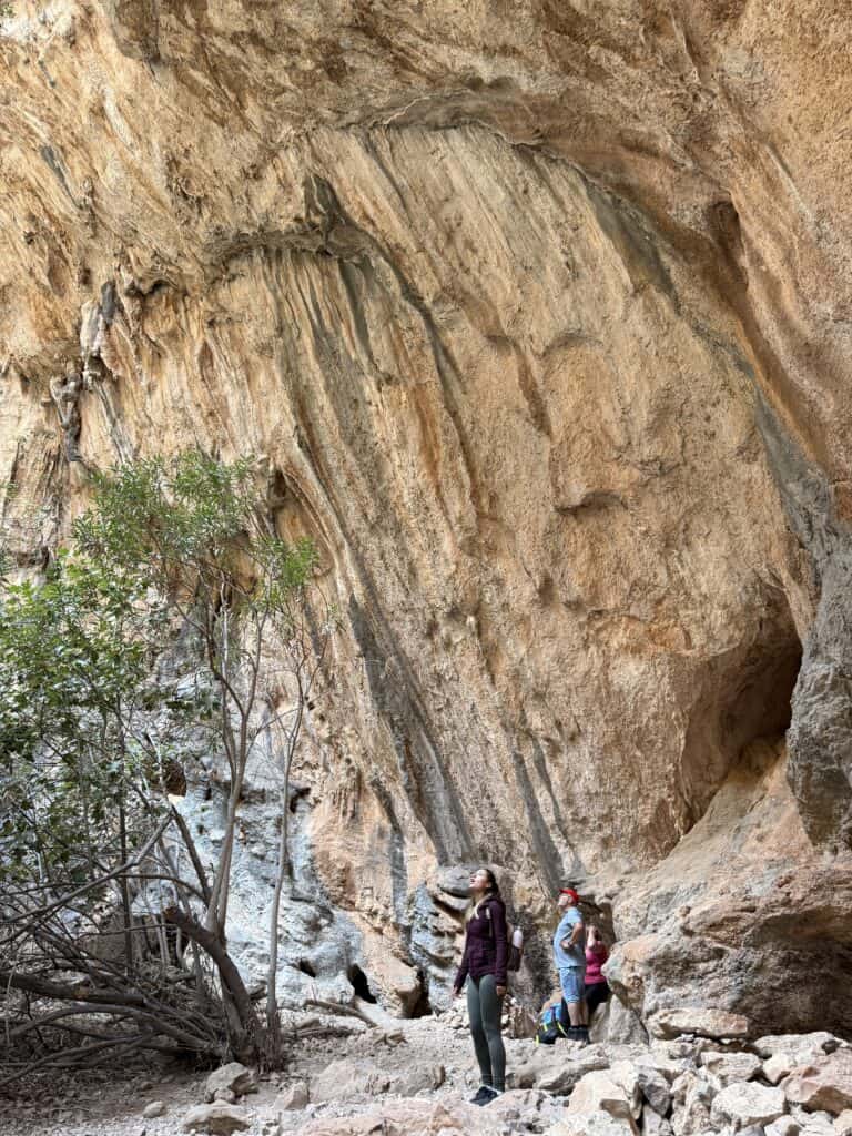 Two people stand at the base of a large, textured rock wall in a rocky outdoor landscape along the Gorropu Gorge hike, with sparse vegetation nearby.