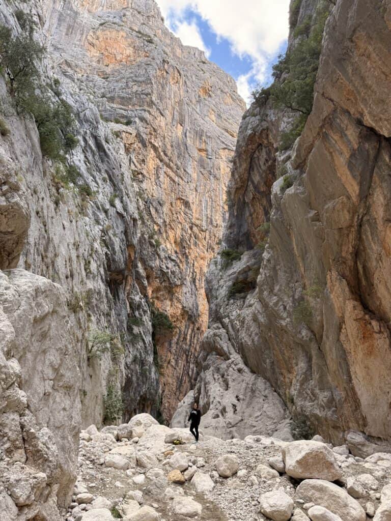 A person stands among large rocks at the base of Su Gorropu Canyon, surrounded by steep, rocky cliffs under a partly cloudy sky.