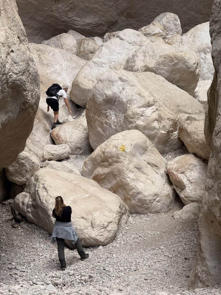 Two people navigate large, light-colored boulders in the stunning Su Gorropu Canyon; one climbs ahead while the other stands below, observing the rugged beauty of this famous Gorropu Gorge hike.