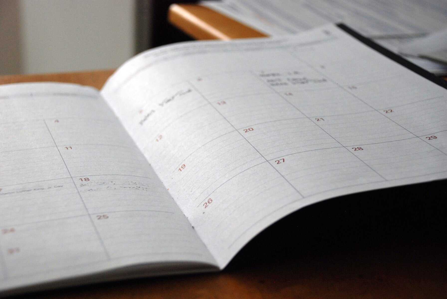 An open desk calendar showing a blank monthly layout with handwritten notes on a few dates, reminders for tasks like comparing Lingoda vs Preply, placed on a wooden surface.