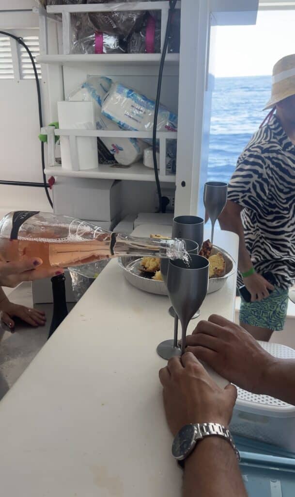 A person pours rosé wine into plastic glasses on a boat near Alghero, Sardinia, with food and another person in a zebra-print shirt nearby. The ocean sparkles in the background—one of many unforgettable things to do in Alghero.
