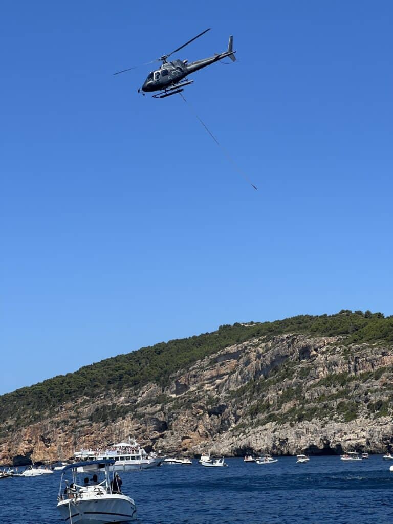 A helicopter with a cable attached flies over a coastal area near Alghero Sardinia, with boats on the water and rocky hills in the background under a clear blue sky—one of the unique things to do in Alghero.