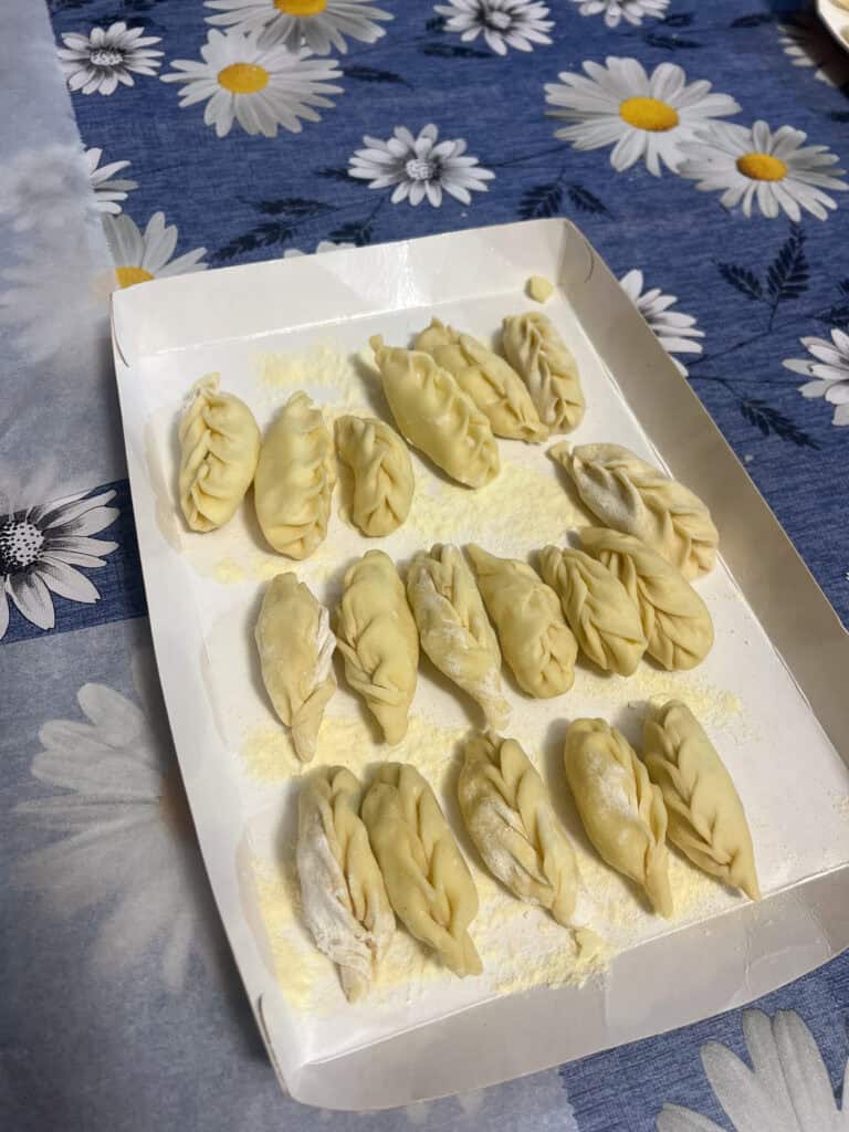 A tray of uncooked, pleated dumplings rests on a dusting of flour, placed on a blue tablecloth with white daisy patterns—perfect inspiration for those exploring Alghero Sardinia and seeking authentic culinary experiences.
