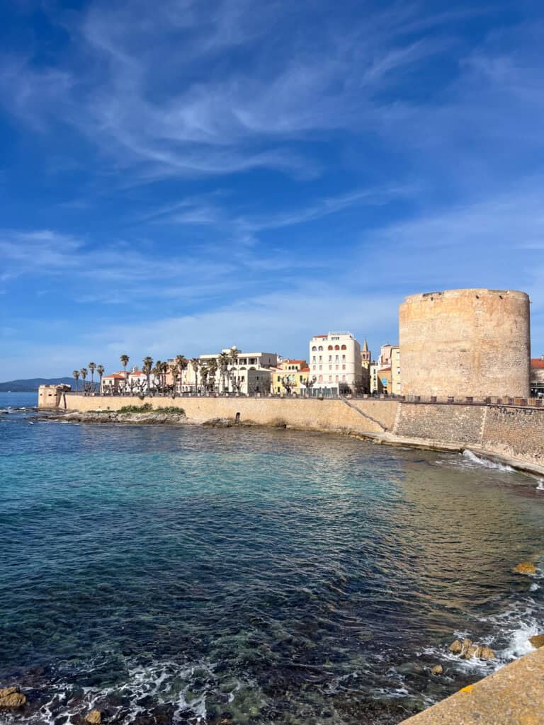 Coastal town of Alghero, Sardinia with a historic stone tower, colorful buildings, palm trees, and clear blue water under a partly cloudy sky—perfect for exploring all the top things to do in Alghero.