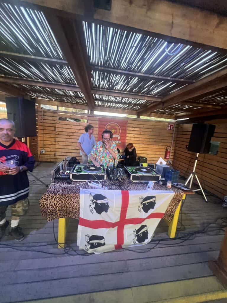 A DJ stands behind turntables on a table draped with a Sardinian flag, performing in an outdoor wooden venue—one of the unique things to do in Alghero, with two other people nearby enjoying the vibrant atmosphere.