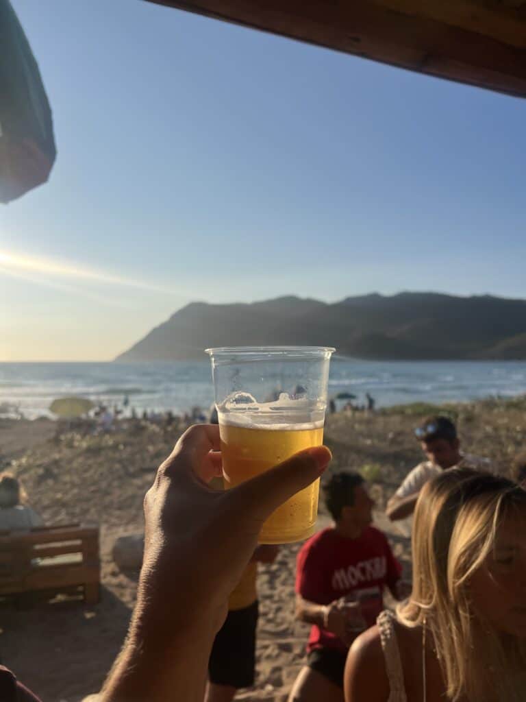 A hand holds a plastic cup of beer up to the sky on a sunny beach in Alghero, Sardinia—one of the top things to do in Alghero—with people, sand, ocean waves, and mountains in the background.