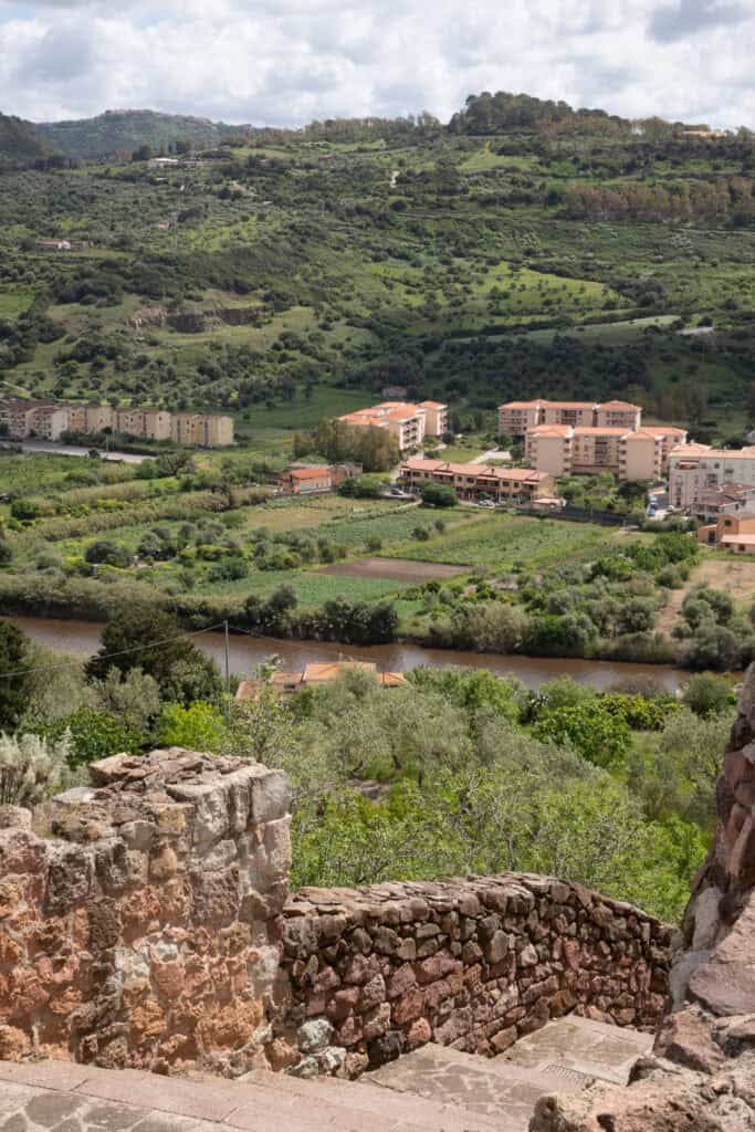 View from stone steps overlooking a river, green fields, and residential buildings with hills and clouds beyond—a peaceful scene that's perfect for exploring while discovering things to do in Alghero Sardinia.