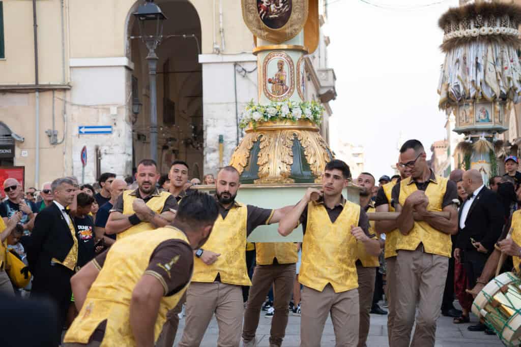 A group of men in matching yellow vests carries a decorated religious float during a street procession, one of the traditional Alghero things to do that immerses you in the culture of Alghero Sardinia, surrounded by fascinated onlookers.