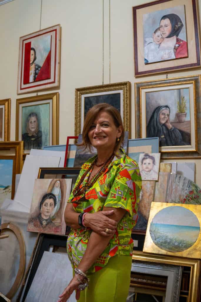 A woman stands smiling in front of a wall and floor filled with framed portraits and landscape paintings in an art studio—one of the inspiring things to do in Alghero, Sardinia.