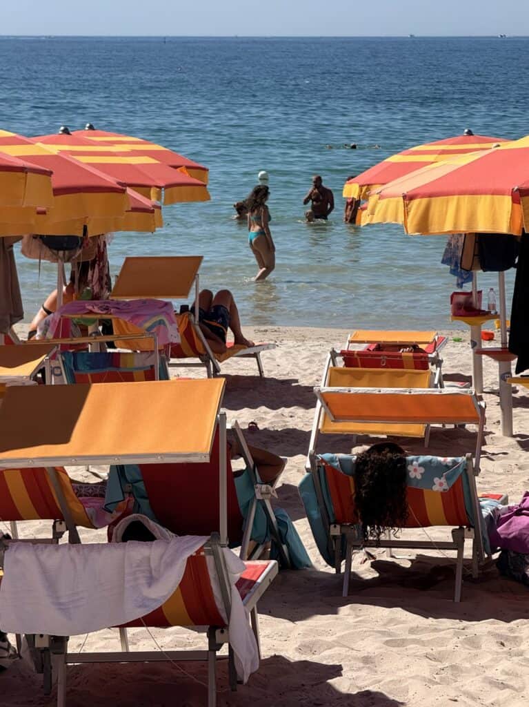 Several people relax on brightly colored orange and red beach chairs under matching umbrellas in Alghero, Sardinia, while others stand in the shallow water—one of the classic things to do in Alghero.